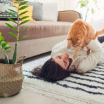 young woman playing with cat on carpet with plant behind them