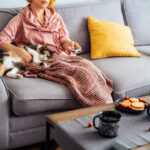 Woman with plaid selecting movie with remote controller and stroking multicolored cat pet on the sofa. Movie night party at home with sweets and hot drinks. Cozy autumn holidays. Selective focus